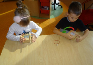 Dwoje dzieci siedzi przy stole. Dziewczynka trzyma w rękach kartonowe pudełeczko z nałożonymi gumkami oraz przełożonym przez nie ołówkiem. Chłopiec zakłada gumki na kartonowe pudełeczko.